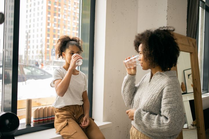 Positive African American mother with daughter in casual wear looking at each other while drinking water from glasses in light room